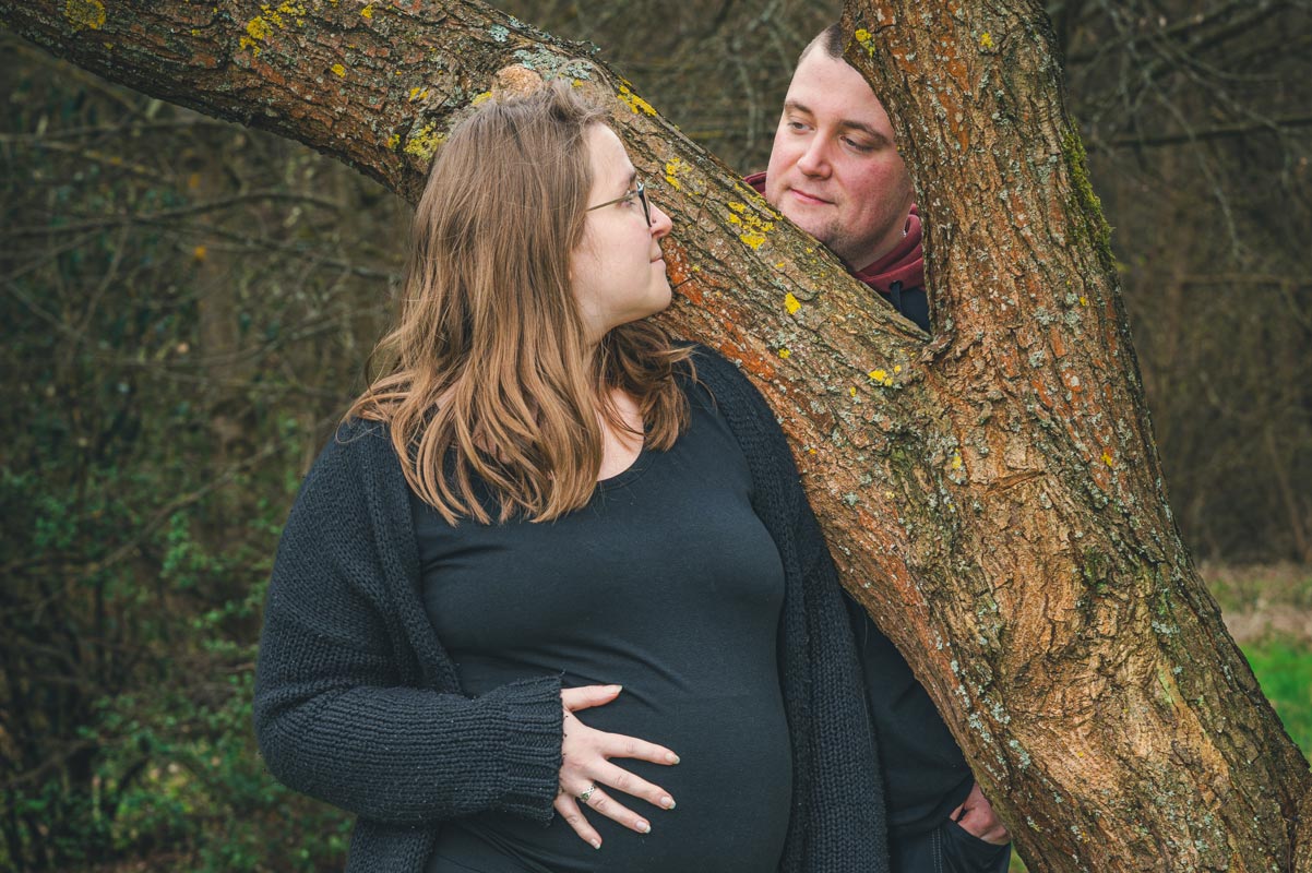 Eine junge schwangere Frau blickt ihren Mann am Baum in die Augen. Die Hand von ihr berührt den Babybauch.