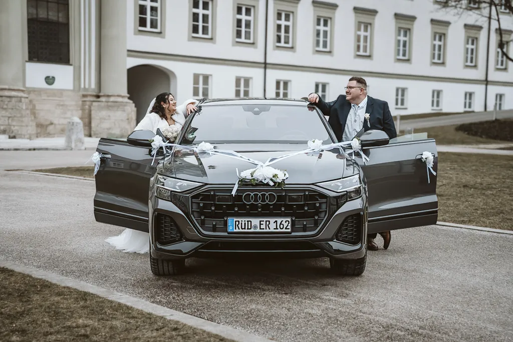 Verbindung über das Autodach hinweg Modernes Brautpaar steht am Hochzeitsauto und hält Blickkontakt über das Dach hinweg. Kreative Bildkomposition beim Paarshooting in Fürstenfeldbruck.