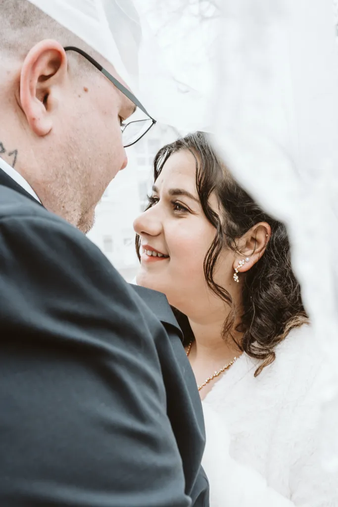 Intimer Moment unter dem Brautschleier Emotionales Hochzeitsfoto: Braut und Bräutigam unter dem Schleier in einer intimen Nahaufnahme mit direktem Augenkontakt.