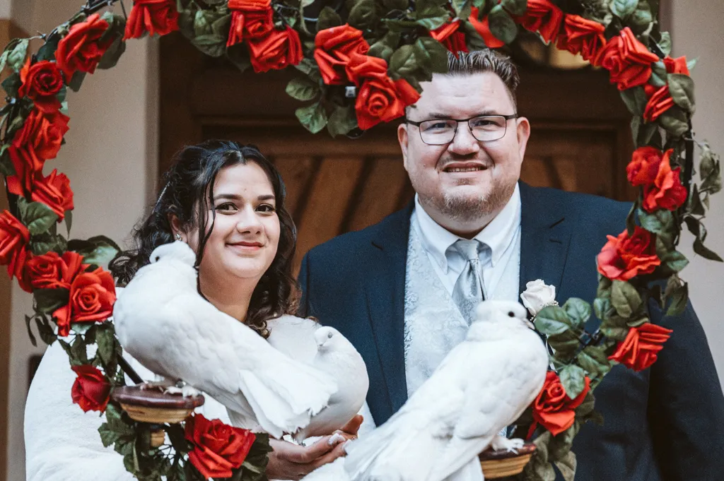 Romantisches Porträt mit Hochzeitstauben und Rosenherz Frisch vermähltes Brautpaar posiert hinter einem Herz aus Rosen mit weißen Hochzeitstauben nach der Trauung in Fürstenfeldbruck.