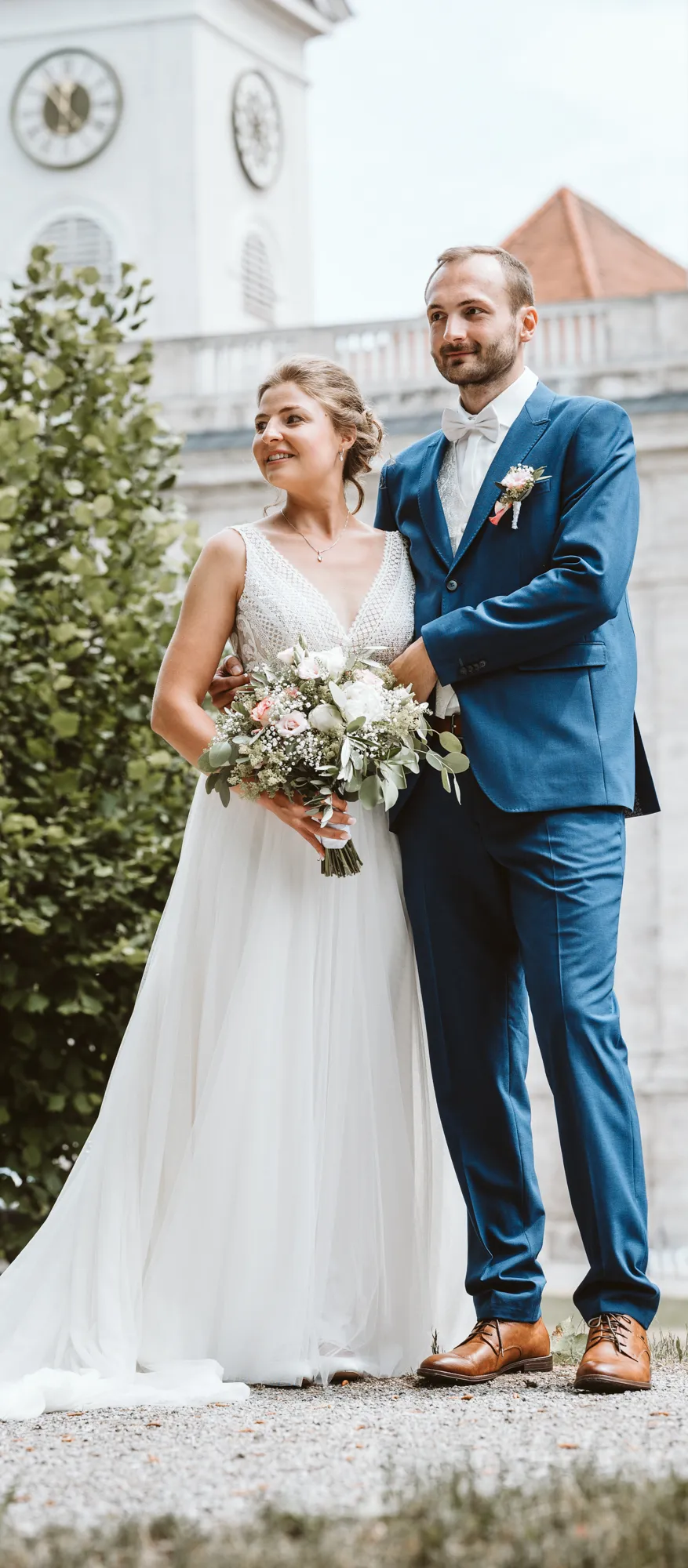 Der Bräutigam im blauen Anzug umarmt seine Braut im weißen Kleid. Beide stehen mit dem Rücken zur Kirche.
