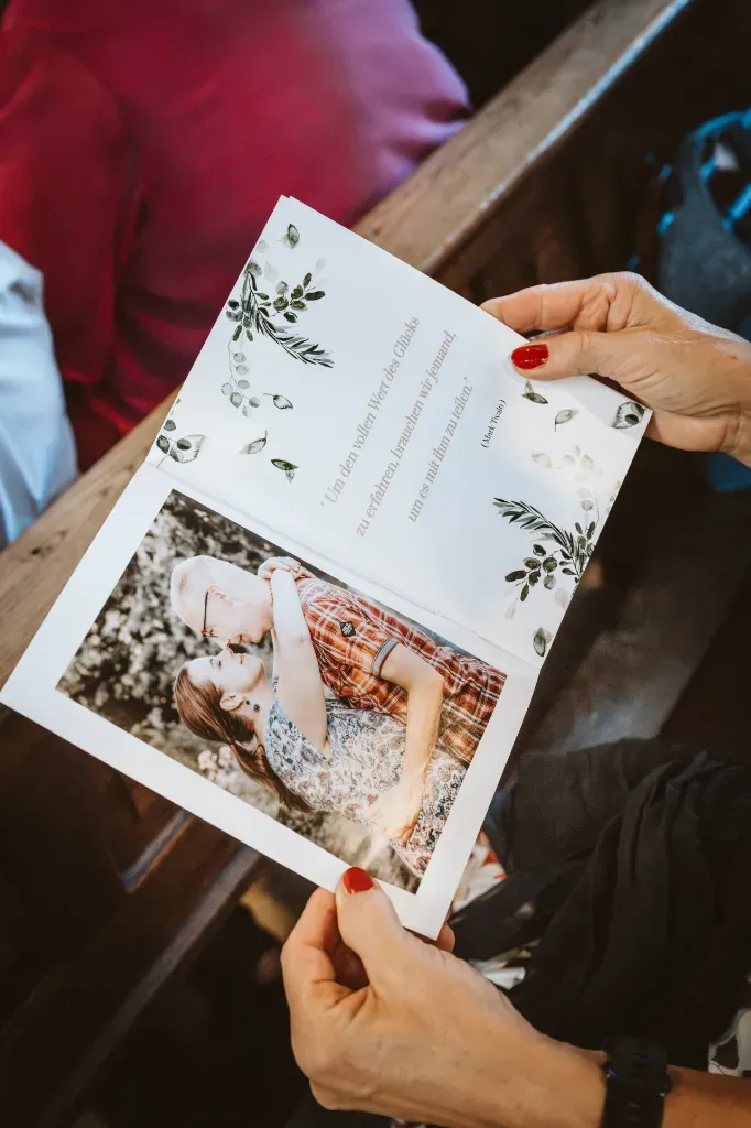 Detail: Hochzeitskarte mit Bild des Brautpaares Hochzeitsreportage Olching: Eine Detailaufnahme zeigt eine Hand, die die Einladungskarte des Brautpaares hält. Auf der Karte ist ein Bild der beiden Verlobten abgebildet.