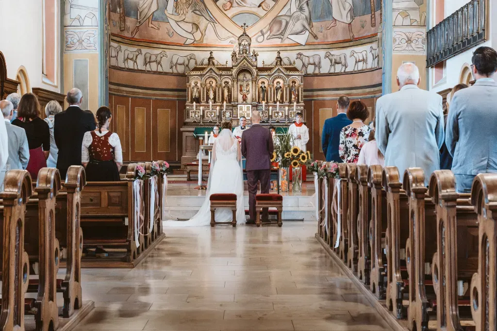 Totale der Zeremonie in der Kirche in Olching Totale aus der Ferne, die das Brautpaar vor dem Altar in der Kirche Olching zeigt. Die Hochzeitsgäste stehen in den Kirchenbänken und folgen der Zeremonie.