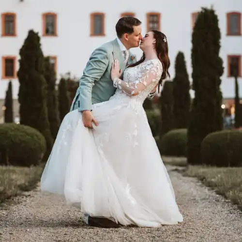 Braut winkt mit Brautstrauß Ich habe das Hochzeitspaar im blauen Oldtimer fotografiert, wobei die Braut den Brautstrauß zum Fenster hinaus hält und winkt. Eine stimmungsvolle Aufnahme von mir als Hochzeitsfotograf Olching, die die freudige Fahrt dokumentiert.
