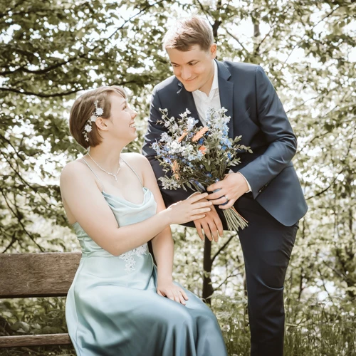 Bräutigam überreicht der Braut Blumen Kreidt Fotografie Olching: Liebevolle Übergabe eines Blumenstraußes vom Bräutigam an seine sitzende Braut.
