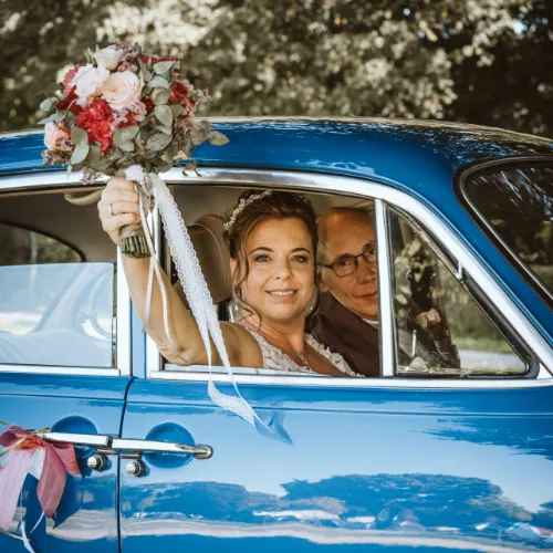 Ich habe das Hochzeitspaar im blauen Oldtimer fotografiert, wobei die Braut den Brautstrauß zum Fenster hinaus hält und winkt. Eine stimmungsvolle Aufnahme von mir als Hochzeitsfotograf Olching, die die freudige Fahrt dokumentiert.
