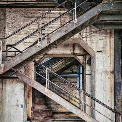 Lostplace einer Fabrikhalle. Eine von der Seite aufgenommene rostige Postesttreppe die zum Obergeschoß führt.